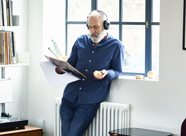 Older man leaning up wall with headphones on and reading a magazine