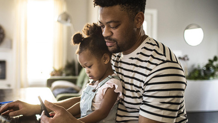 Man with young child on lap, looking at phone and laptop 